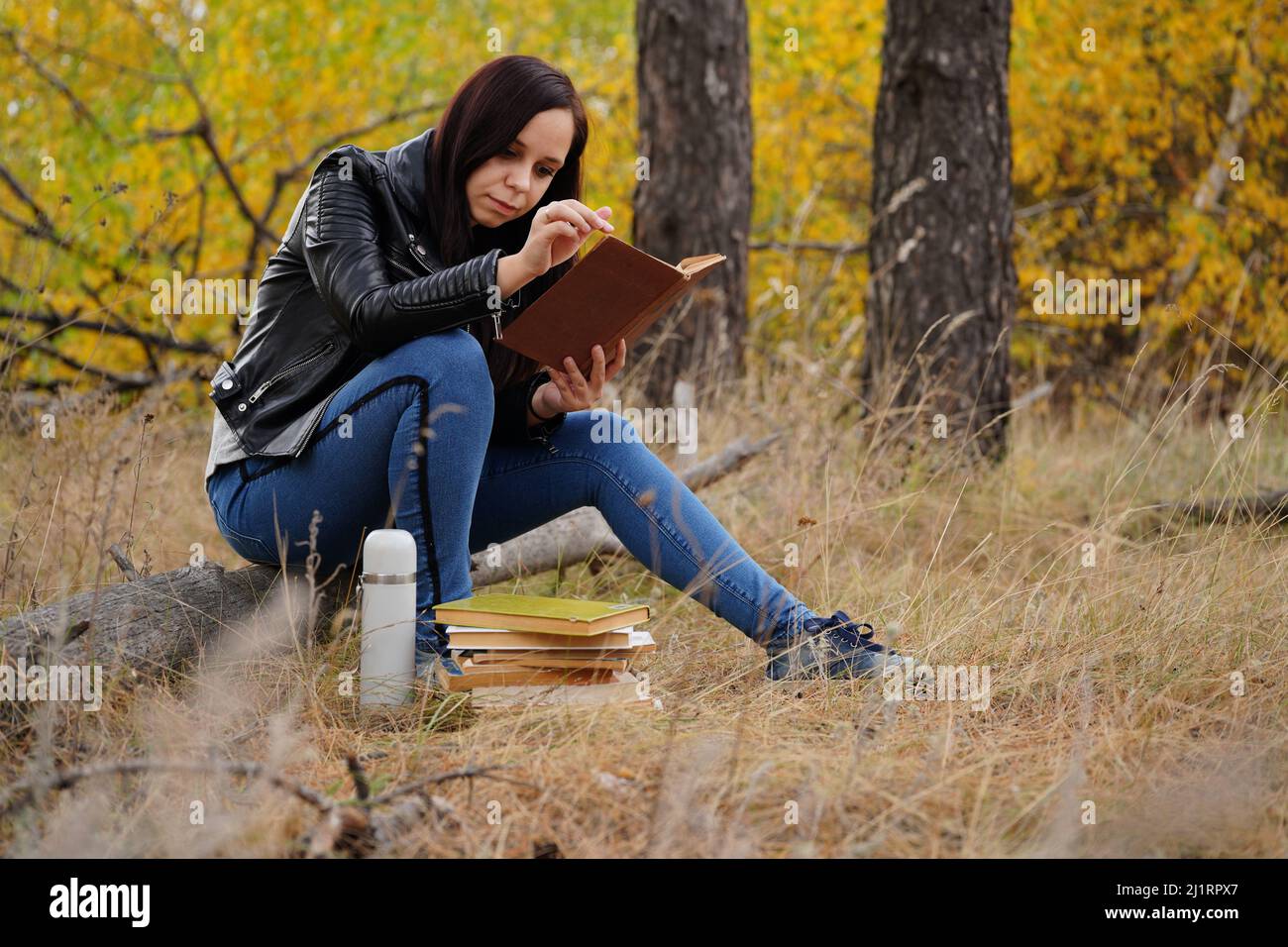 Eine junge, schöne Frau mit langen, dunklen Haaren in legerer Kleidung sitzt auf einem Holzbuch und liest ein Buch Stockfoto