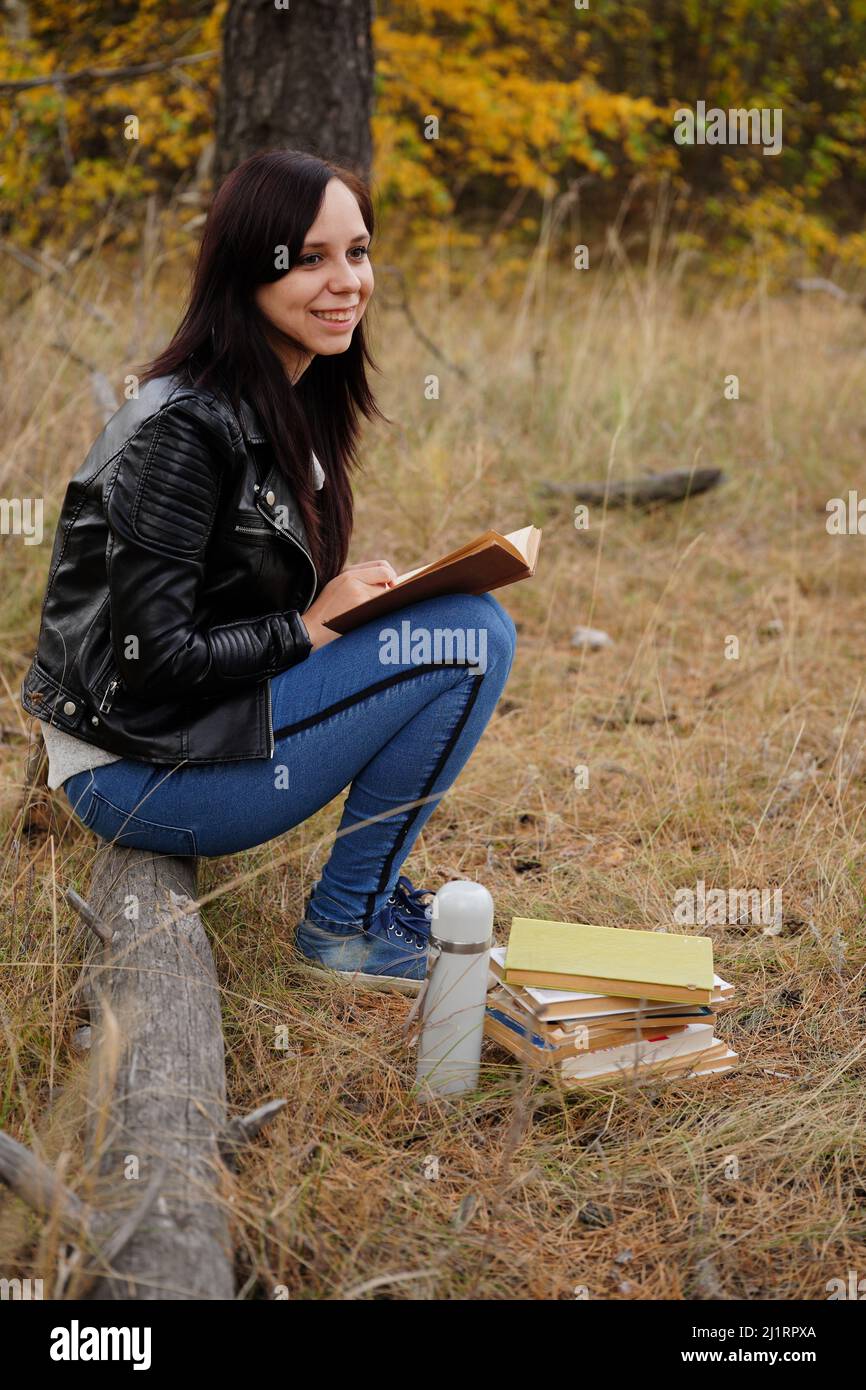 Eine junge, schöne Frau mit langen, dunklen Haaren in legerer Kleidung sitzt auf einem Holzbuch und liest ein Buch Stockfoto