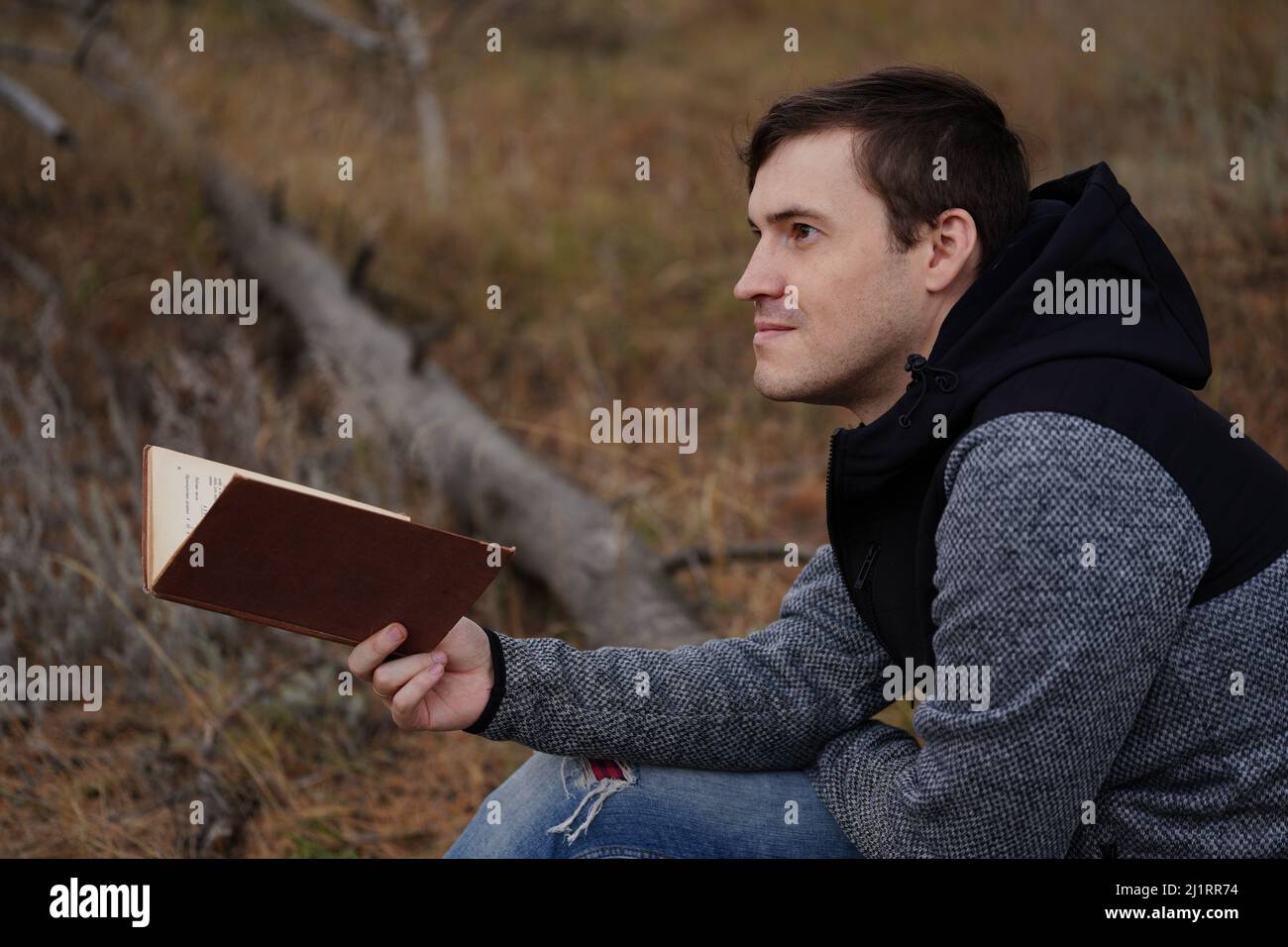 Ein junger, gutaussehender Mann in lässiger Kleidung sitzt auf einem Holzbuch und liest im Herbstwald ein Buch Stockfoto