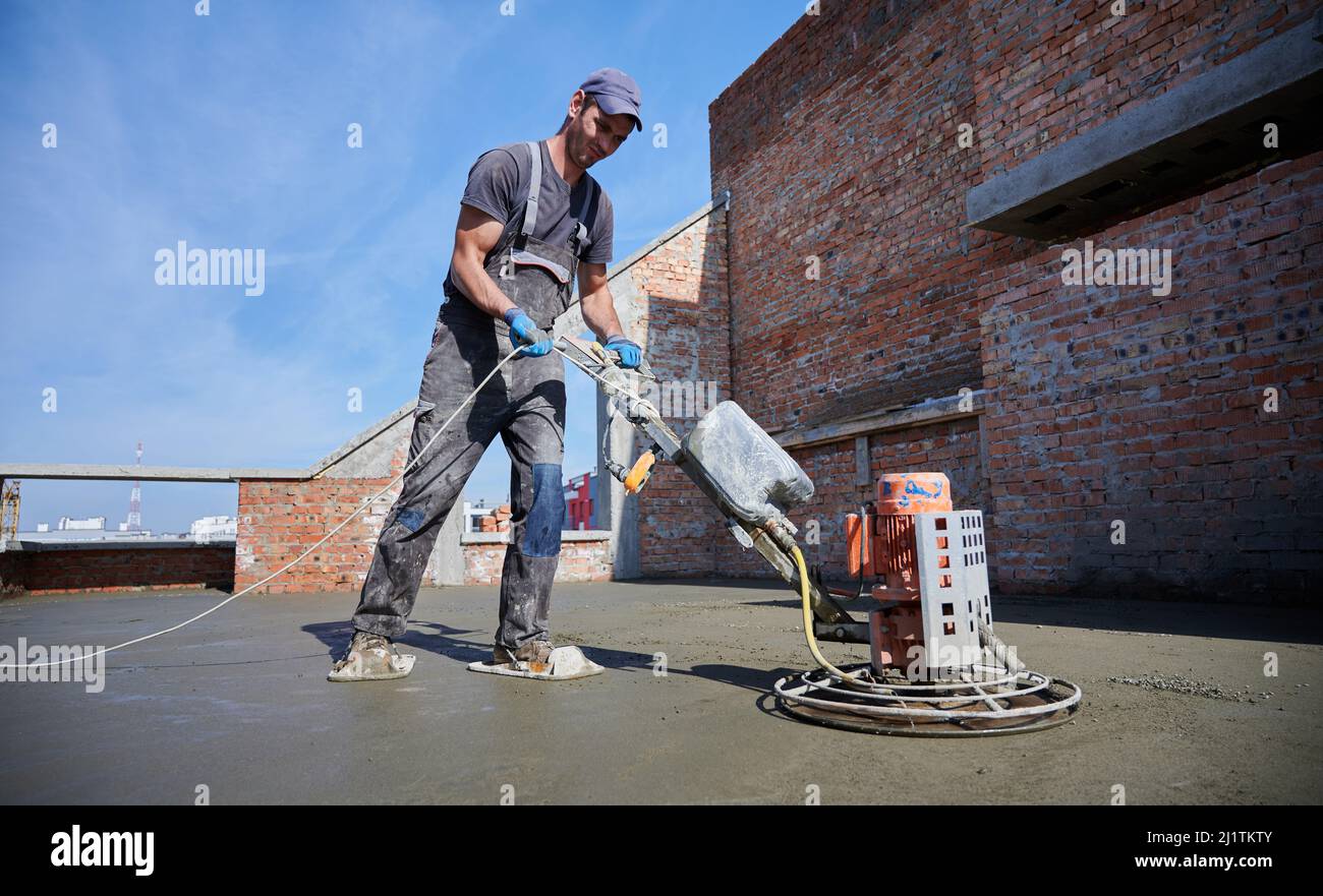Junger Baumeister in Arbeitskleidung, der mit einer Fugenschleifmaschine für die Ausrichtung und Glättung der Bodenoberfläche arbeitet. Vor dem Auftragen von Deckbelägen den Boden verfugen. Stockfoto