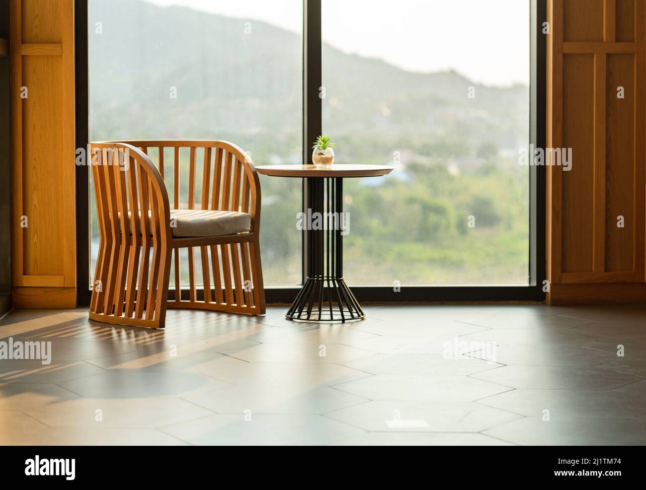 Schöner Holzsessel und runder Tisch mit kleinem Glas-Pflanzentopf, alle zusammen am Spiegel großes Fenster gegen Abendsonne, leer Stockfoto