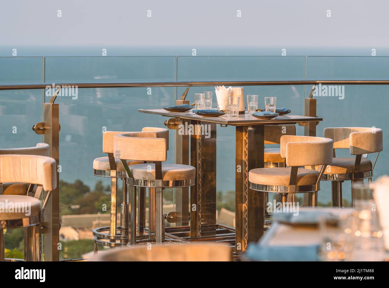 Stilvoller Tisch und Hocker eines Restaurants im Freien auf dem Dach eines Gebäudes, Geschirr auf dem Tisch, Glaszaun, verschwommener Hintergrund des Himmels Stockfoto