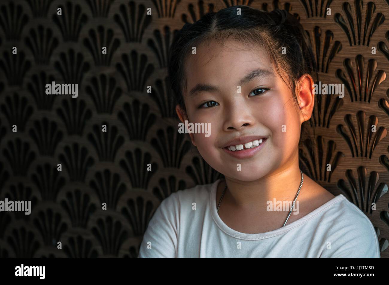 Portrait von asiatischen kleinen Mädchen mit süßen Lächeln, Augen suchen Kamera, Hintergrund der schönen Dekoration Wand, natürliches Sonnenlicht aus dem Fenster. Asiatischer Kanal Stockfoto