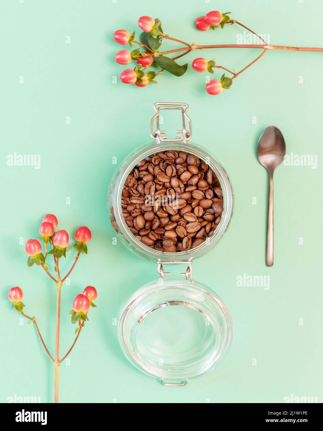 Bio-Kaffeebohnen in einem Glas mit Löffel und roten Blumen auf Minze Hintergrund. Umweltfreundliche Zusammensetzung mit natürlichem Robusta. Kaffee zubereiten. Fla Stockfoto