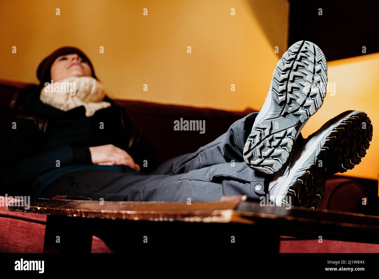 Frau in warmen Kleidern sitzt auf der Couch und legt die Füße in Schuhe auf den Tisch, während sie darauf wartet, nach draußen zu gehen Stockfoto