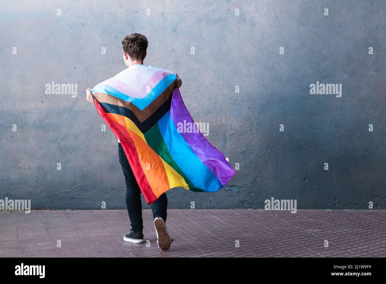 Ganzer Körper einer anonymen Person, die eine Regenbogenflagge und Transgender-Farben in erhobenen Armen hält, während sie gegen eine graue Wand springt Stockfoto