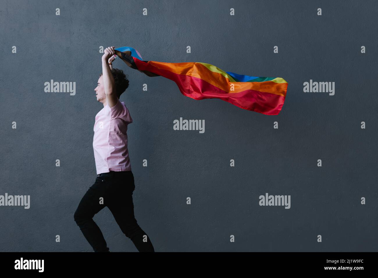 Seitenansicht einer Person, die eine Regenbogenflagge und Transgender-Farben in erhobenen Armen hält, während sie gegen eine graue Wand springt Stockfoto