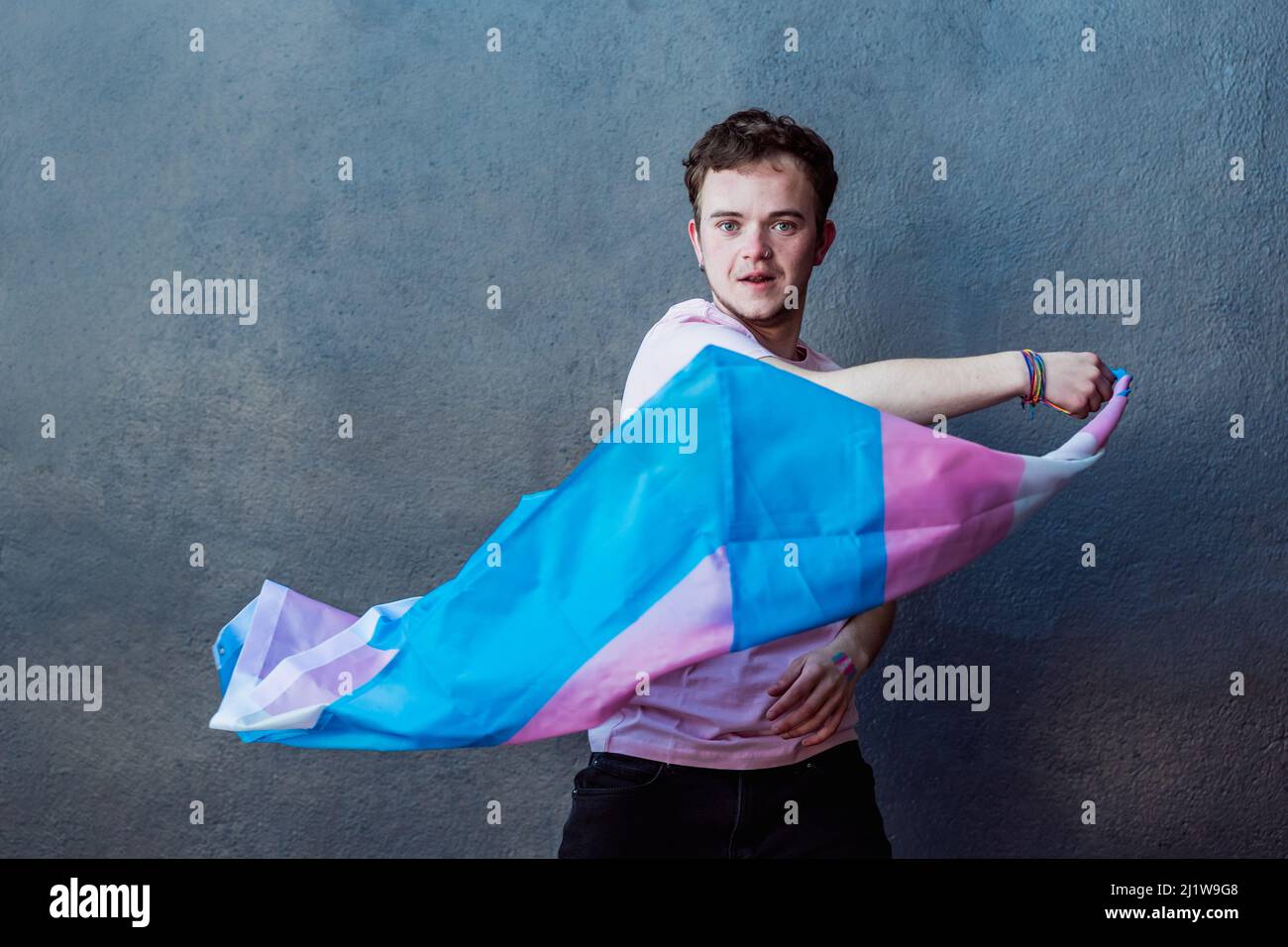 Mann mit farbenfroher Transgender-Flagge mit Streifen, während er auf grauem Hintergrund im hellen Raum steht Stockfoto