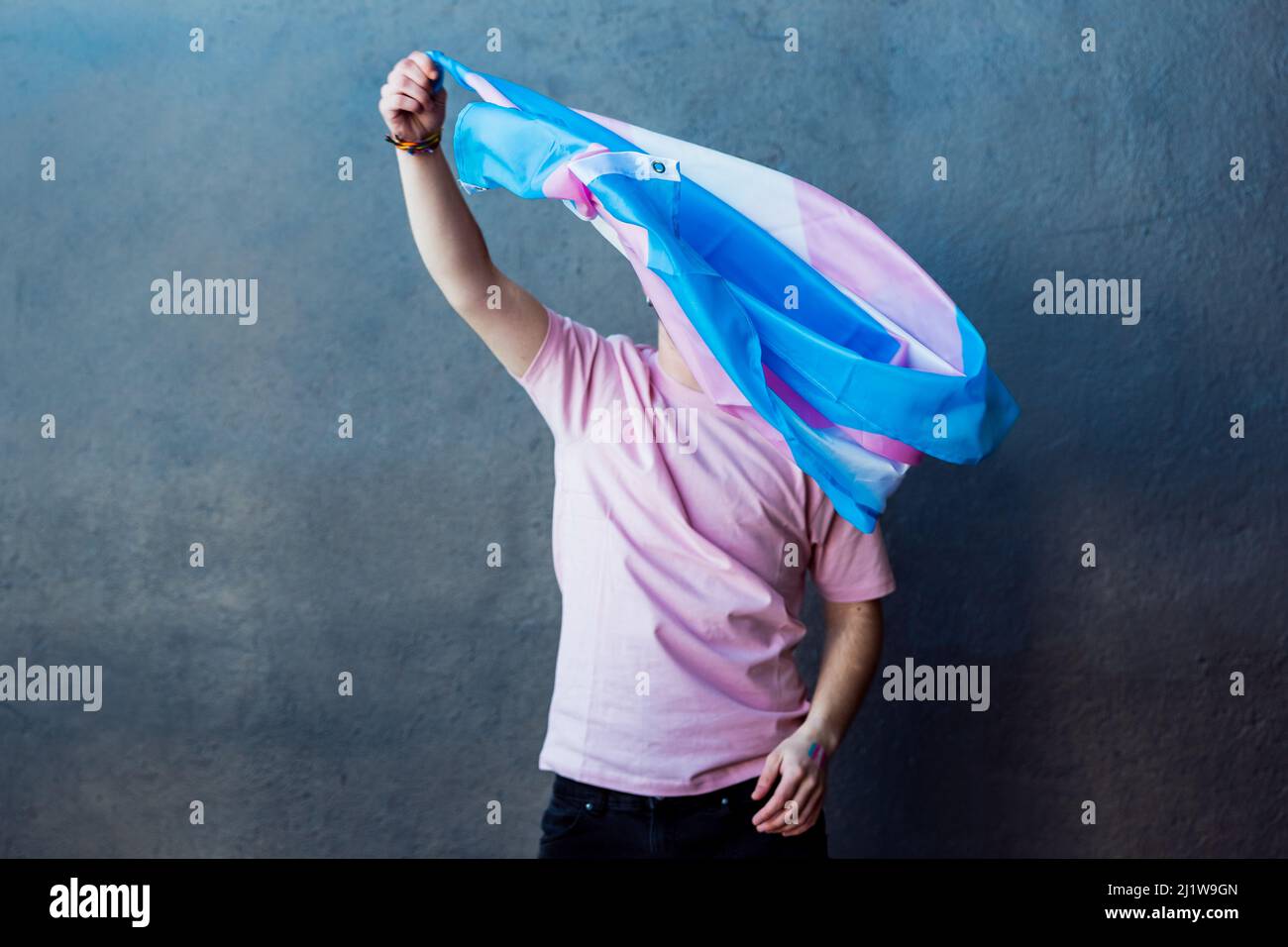 Anonymer Mann mit farbenfroher Transgender-Flagge mit Streifen, während er auf grauem Hintergrund in einem hellen Raum steht Stockfoto