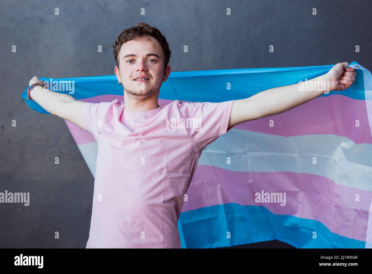 Mann mit farbenfroher Transgender-Flagge mit Streifen, während er auf grauem Hintergrund im hellen Raum steht Stockfoto