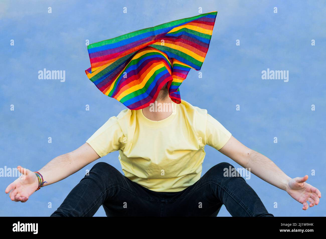 Nicht erkennbare Person, die Arme und Beine mit farbenfroher Regenbogenfahne auf dem Kopf ausbreitet, während sie vor blauem Hintergrund sitzt Stockfoto