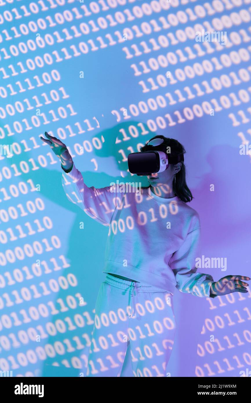 Frau in legerer Kleidung und moderne schwarze VR-Brille mit verschiedenen projizierten Zahlen, während sie den Cyberspace im Zimmer erkundet Stockfoto