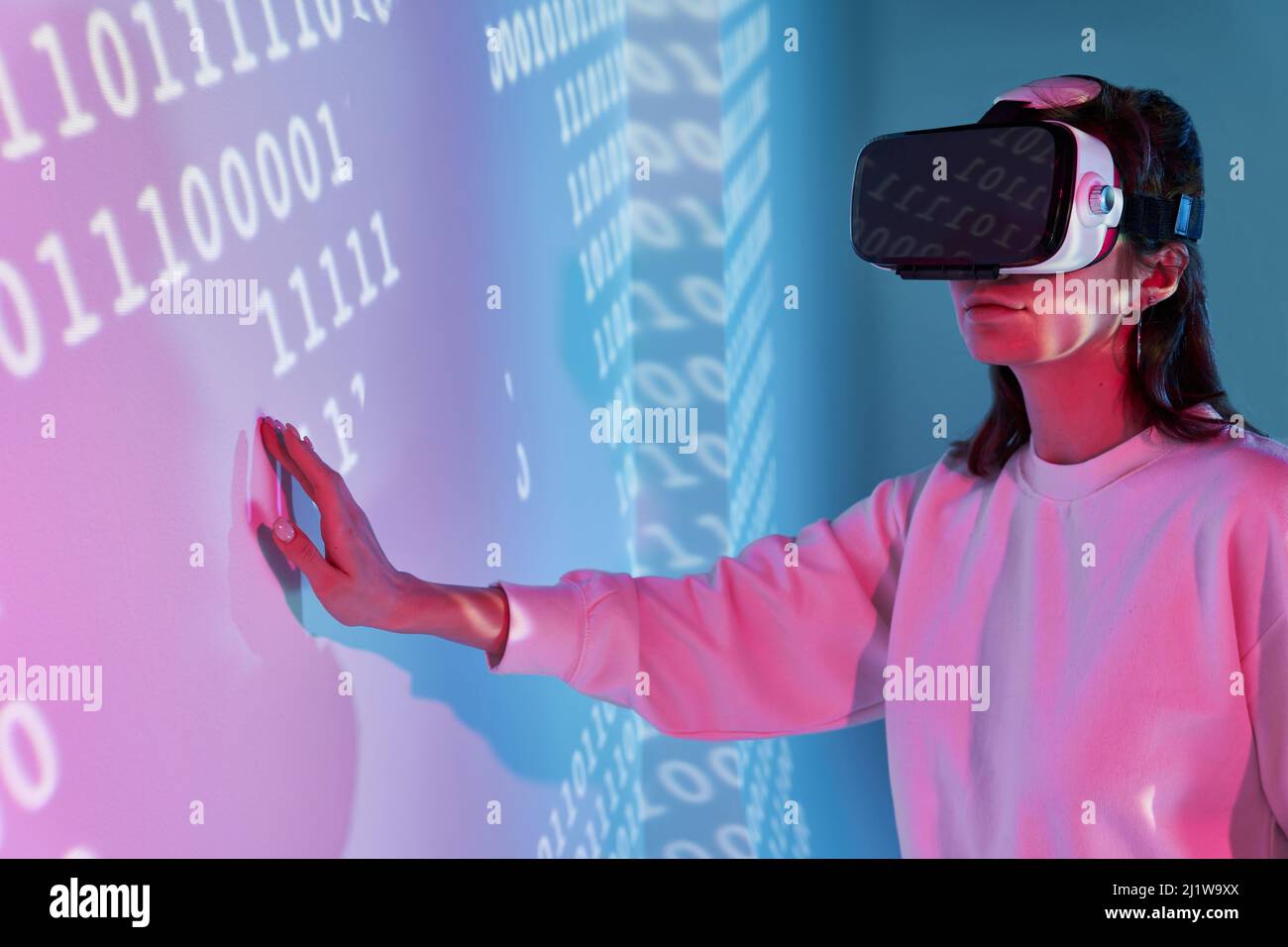 Frau in legerer Kleidung und moderne schwarze VR-Brille berühren die Wand mit verschiedenen projizierten Zahlen, während sie den Cyberspace im Zimmer erkunden Stockfoto