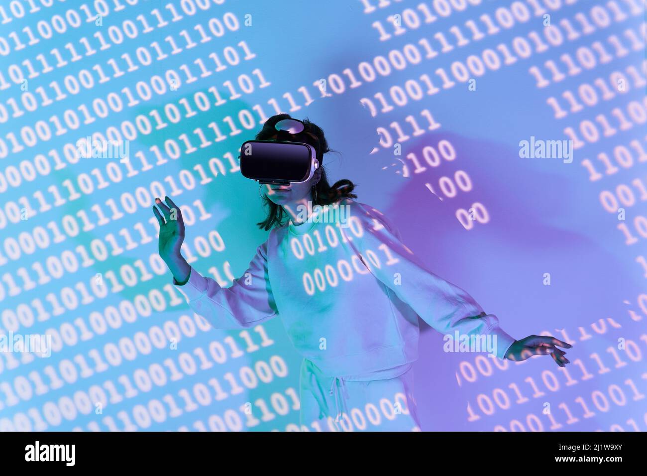 Frau in legerer Kleidung und moderne schwarze VR-Brille mit verschiedenen projizierten Zahlen, während sie den Cyberspace im Zimmer erkundet Stockfoto