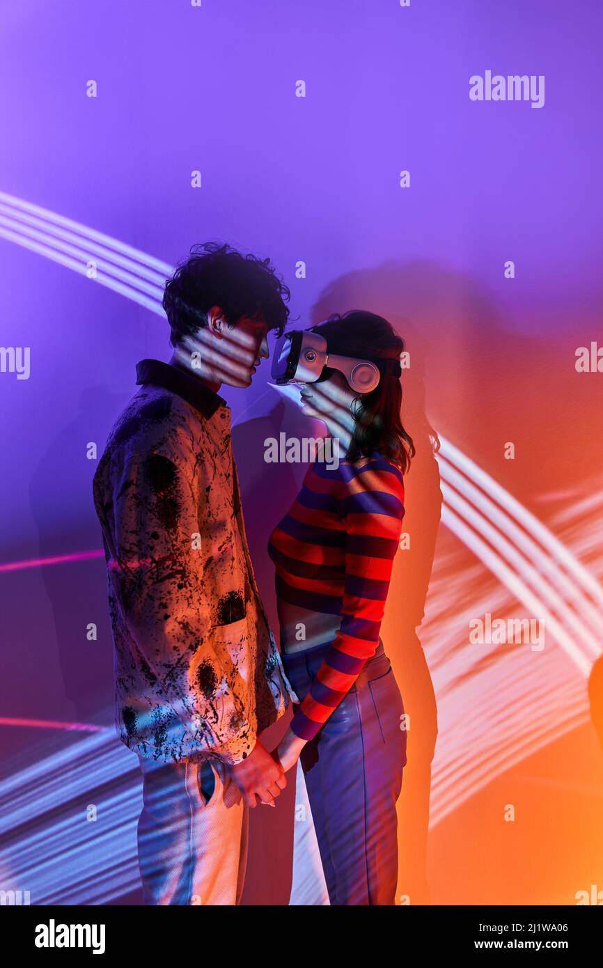 Seitenansicht eines Mannes, der die Hände einer Freundin in einer modernen VR-Brille hält und an der Wand mit leuchtenden Lichtern im Studio steht Stockfoto