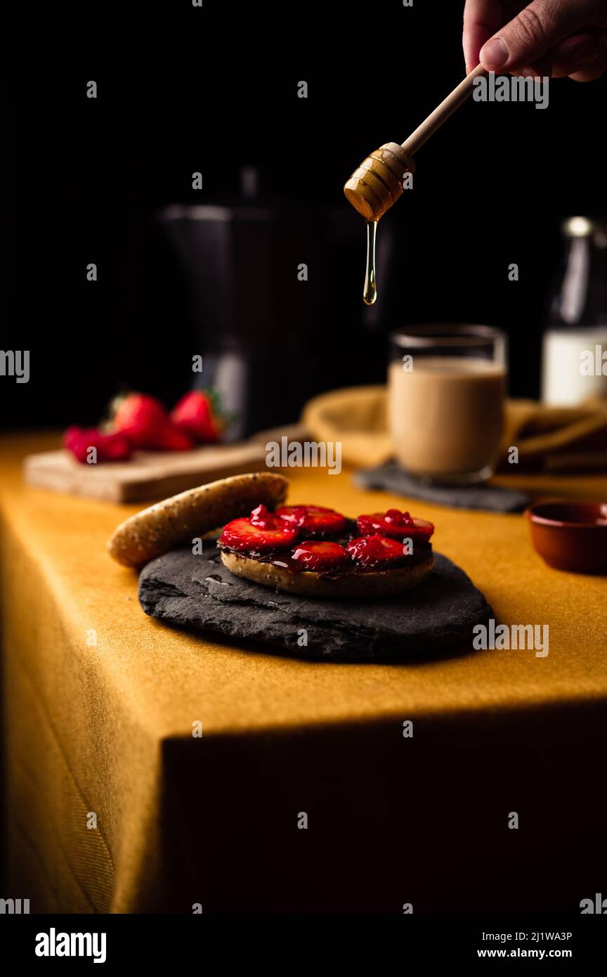 Crop anonyme Person schmieren Honig auf Bagel mit frischen Erdbeeren und Schokoladenpaste zum Frühstück mit frischem Kaffee aus Moka Topf Stockfoto