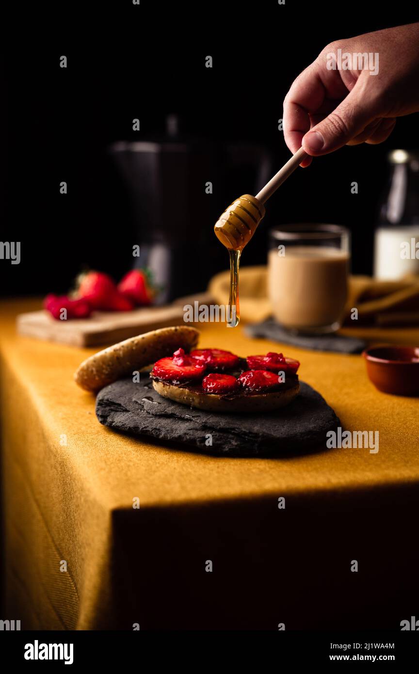 Crop anonyme Person schmieren Honig auf Bagel mit frischen Erdbeeren und Schokoladenpaste zum Frühstück mit frischem Kaffee aus Moka Topf Stockfoto