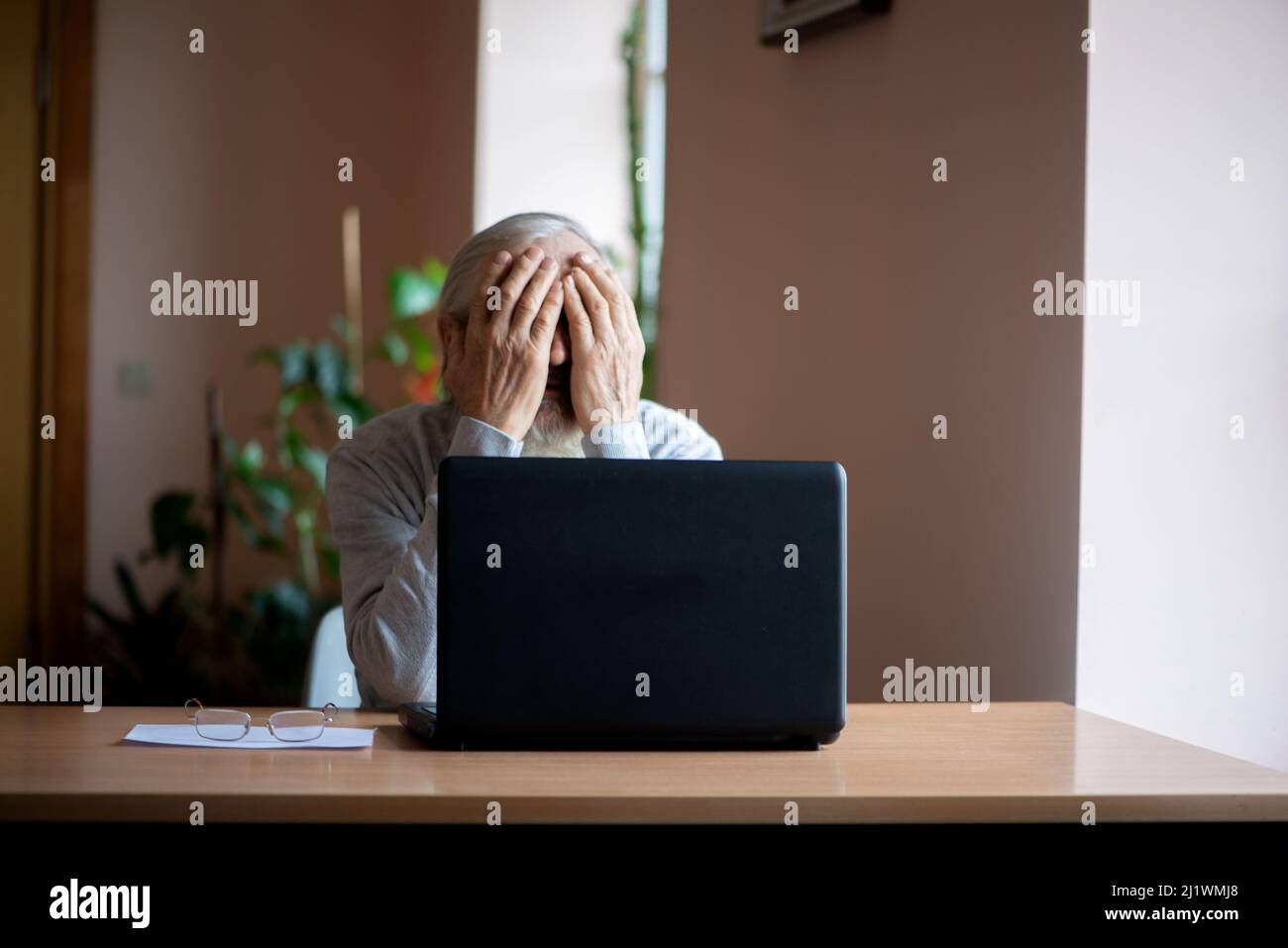 Ein müder, gebrochener, resigniert, beschäftigt älterer Mann sitzt vor einem Laptop, Computer und bedeckt sein Gesicht mit der Hand. Der traurige Senior überprüft seine Klimaanlage Stockfoto