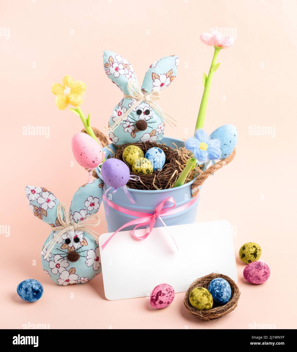 Niedliche Kinder-Osterkarte mit kleinem blauen Eimer, Nester, bunten Wachteleiern, dekorativen Eiern, gestrickten Farbblumen, Stoffhasen, leere Notiz auf pa Stockfoto