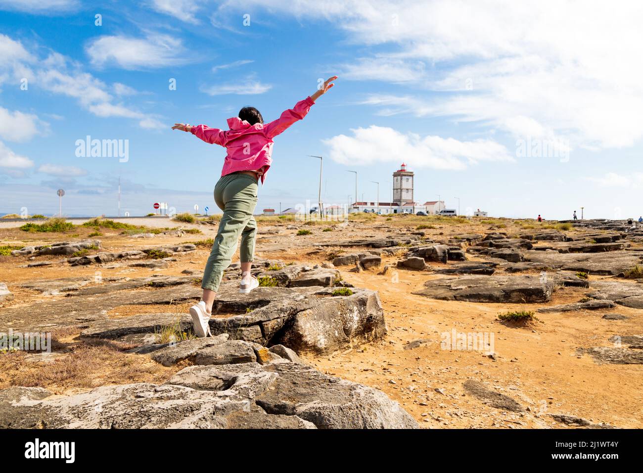 Mädchen springt auf Felsen mit ausgestreckten Armen im Kap Carvoeiro mit dem Leuchtturm im Hintergrund Stockfoto