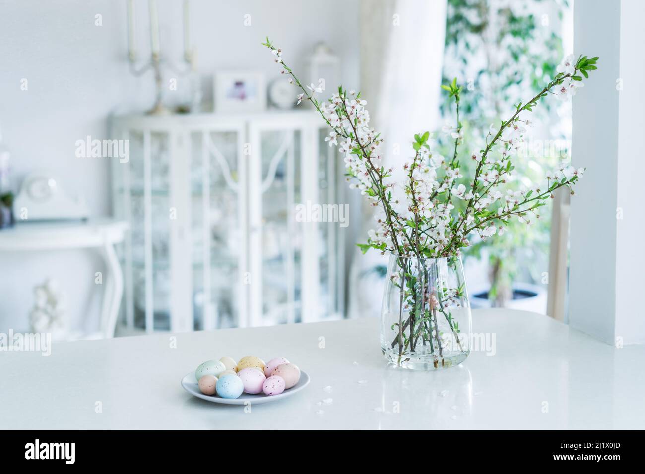 Blühende Baumzweige in der Vase und farbige ostereier auf dem Teller auf weißem Küchentisch mit klassischem Innenhintergrund. Osterfest Stockfoto