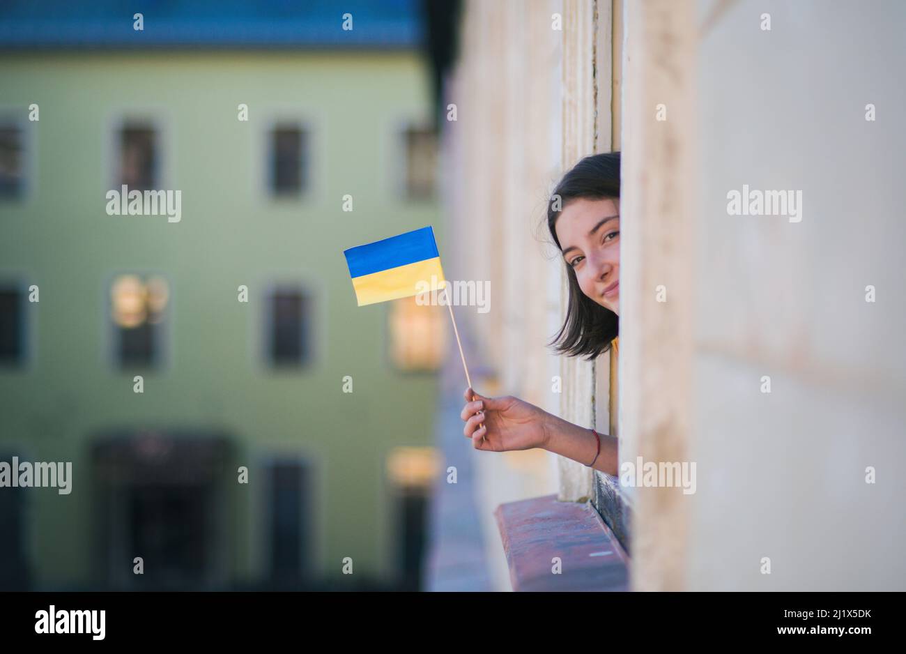 Glückliches Mädchen, das die ukrainische Flagge aus dem Fenster ragt und die Kamera anschaut, Solidarität mit der Ukraine im Kriegskonzept. Stockfoto