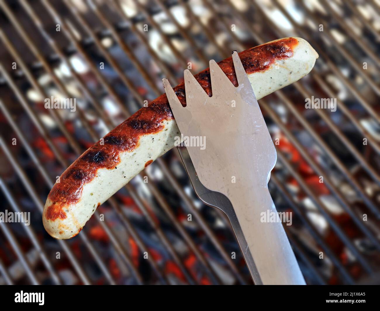 Edelstahl-tong mit Bratwurst, deutsche Wurst über dem bbq-Grill mit glühender Holzkohle und Grillrost im Hintergrund Stockfoto