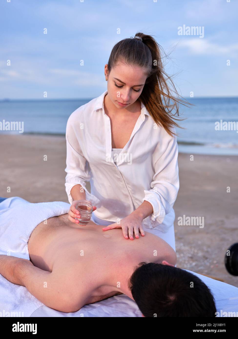 Ein junger Chiromassage-Therapeut gießt einem jungen Mann auf einem Massageliege am Strand von Valencia Öl auf den Rücken. Stockfoto