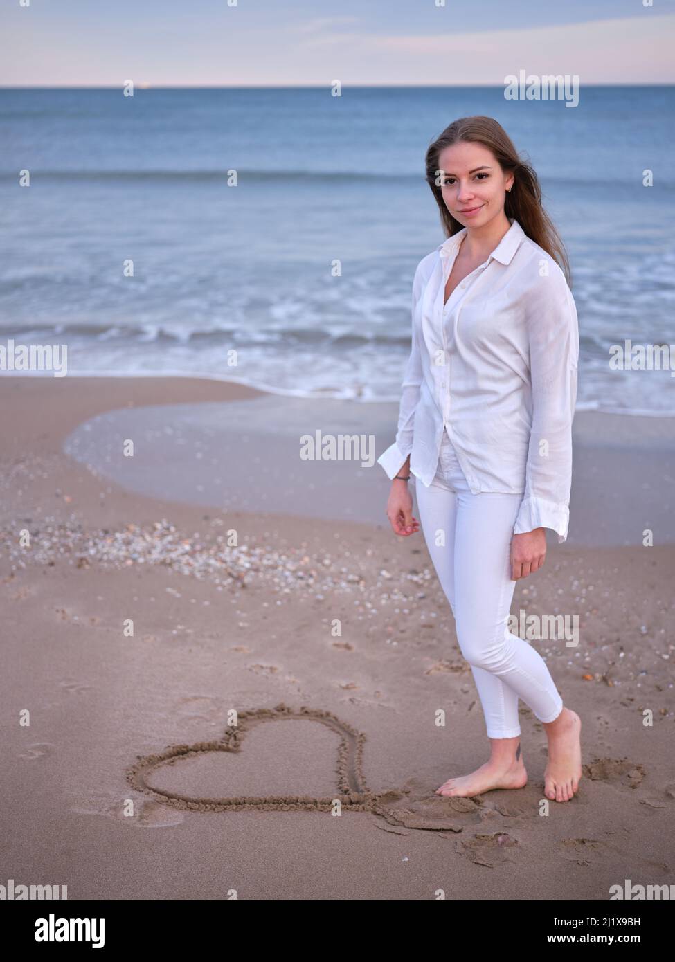 Eine junge Frau in Weiß, die am Strand steht, mit einem im Sand gezeichneten Herzen und der Kamera zuschaut. Stockfoto
