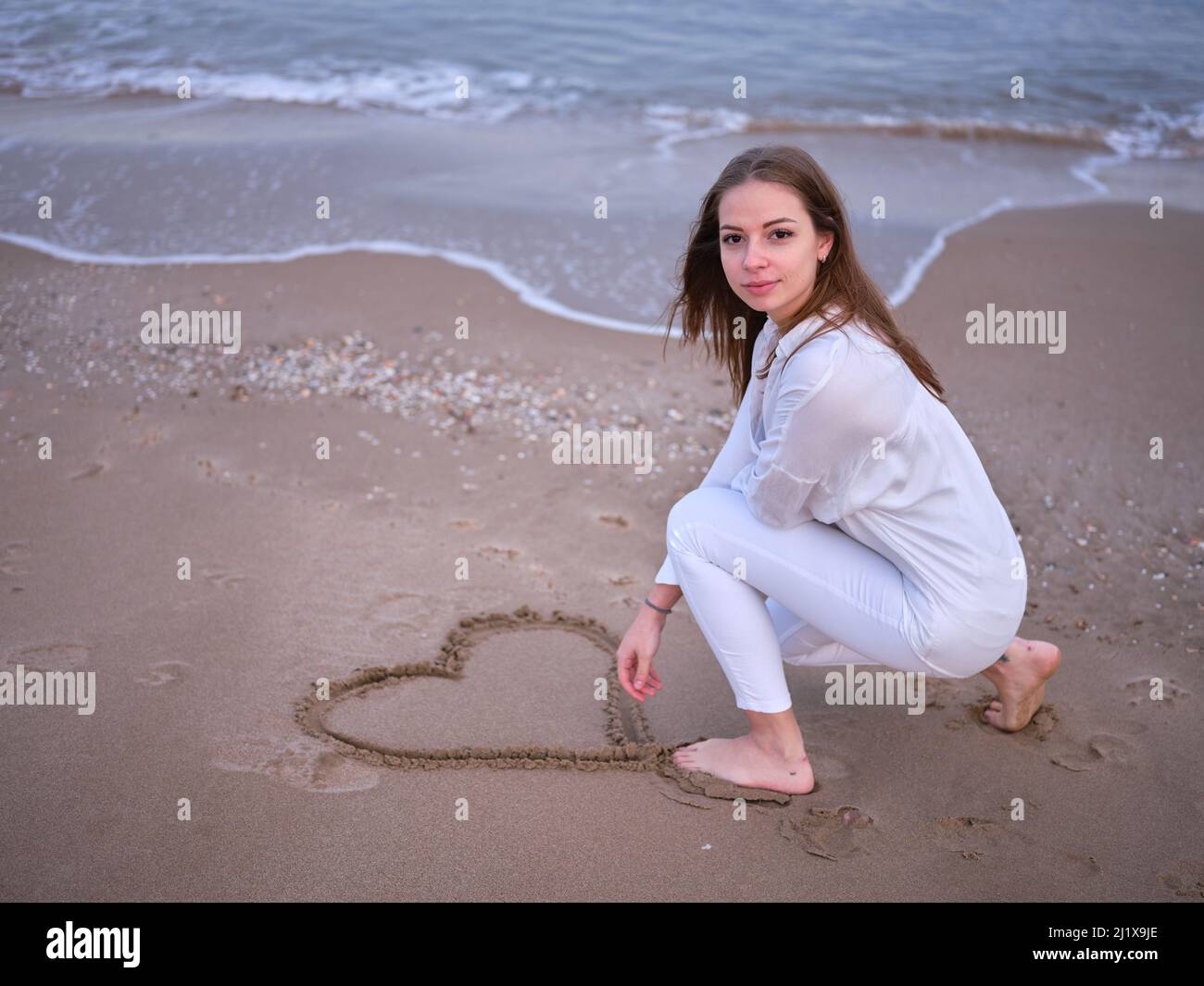 Eine junge Frau in Weiß am Strand, die ein Herz in den Sand zieht und auf die Kamera schaut. Stockfoto