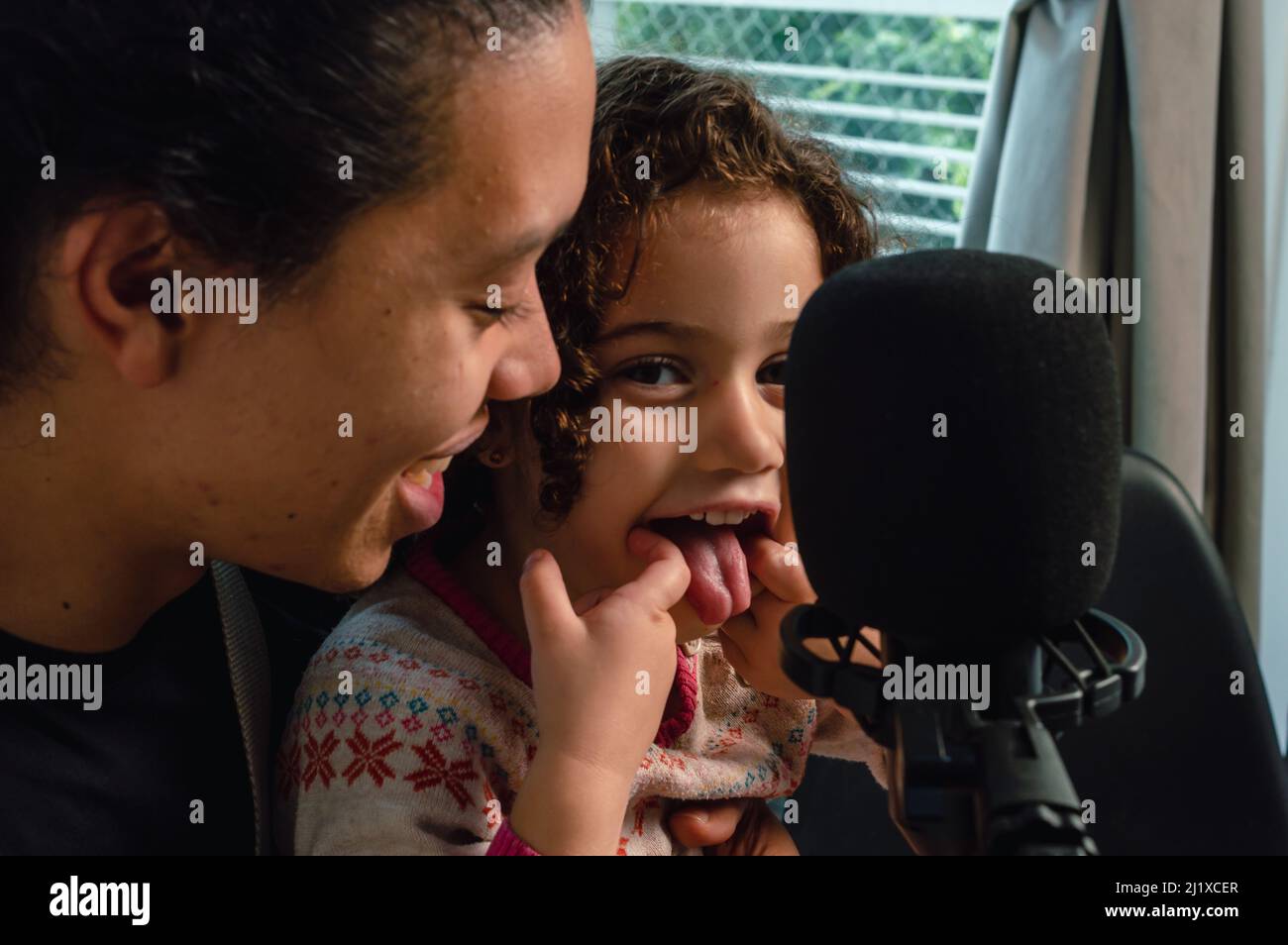 Nahaufnahme kleines kaukasisches Mädchen, das aus der Zunge ragt und glücklich mit ihrem Vater im Musikstudio auf die Kamera blickt. Musik machen und zusammen spielen Stockfoto