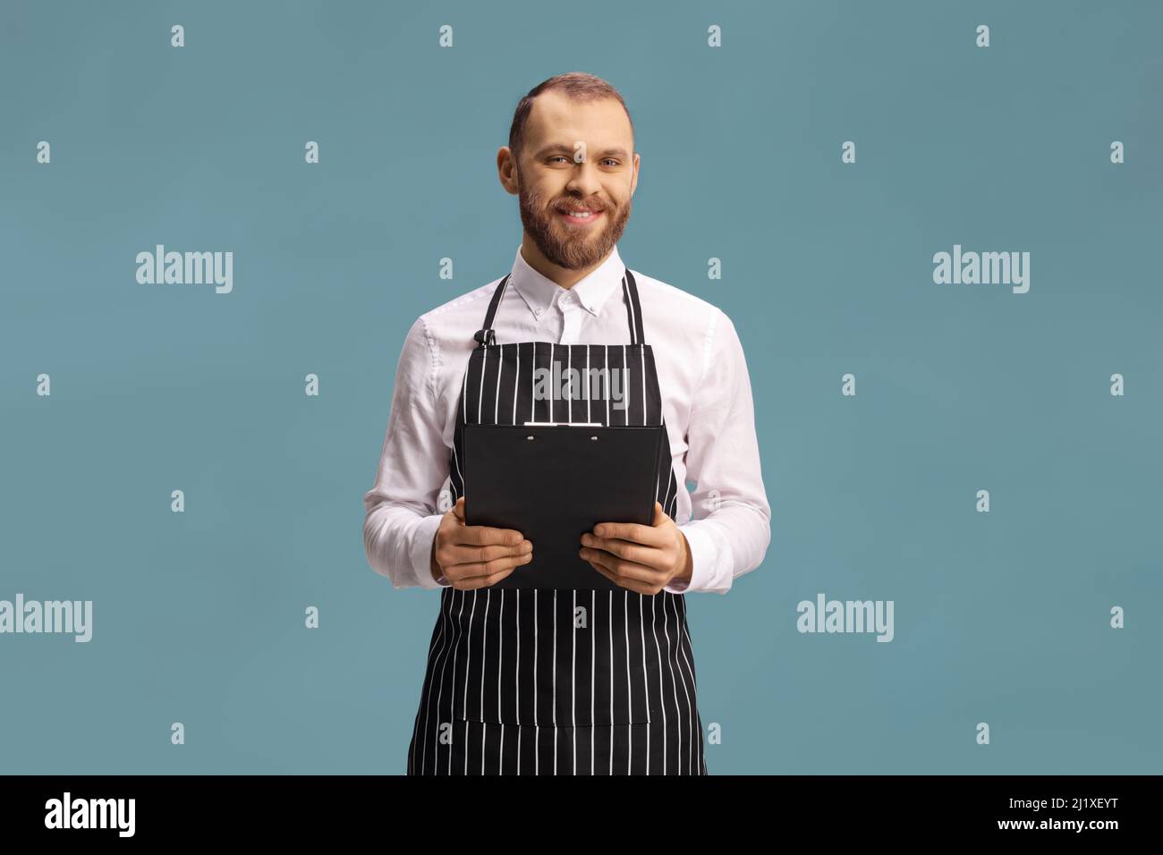 Kellner trägt eine Schürze und hält ein Menü isoliert auf blauem Hintergrund Stockfoto