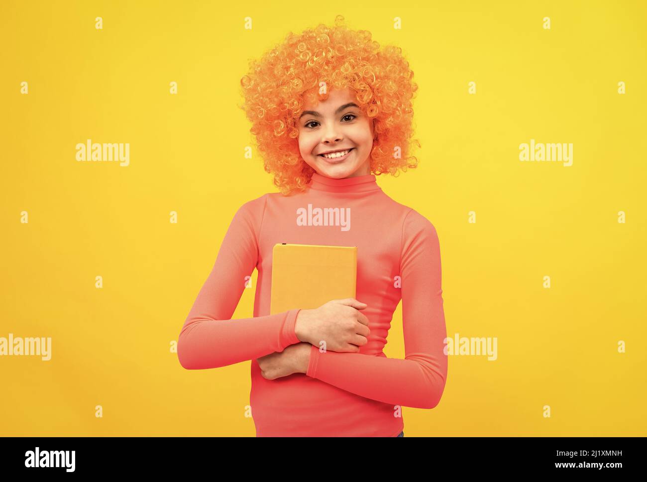 Glückliches Mädchen Kind mit orangen Haaren in rosa Poloneck Lächeln halten Buch gelben Hintergrund, Schule Stockfoto