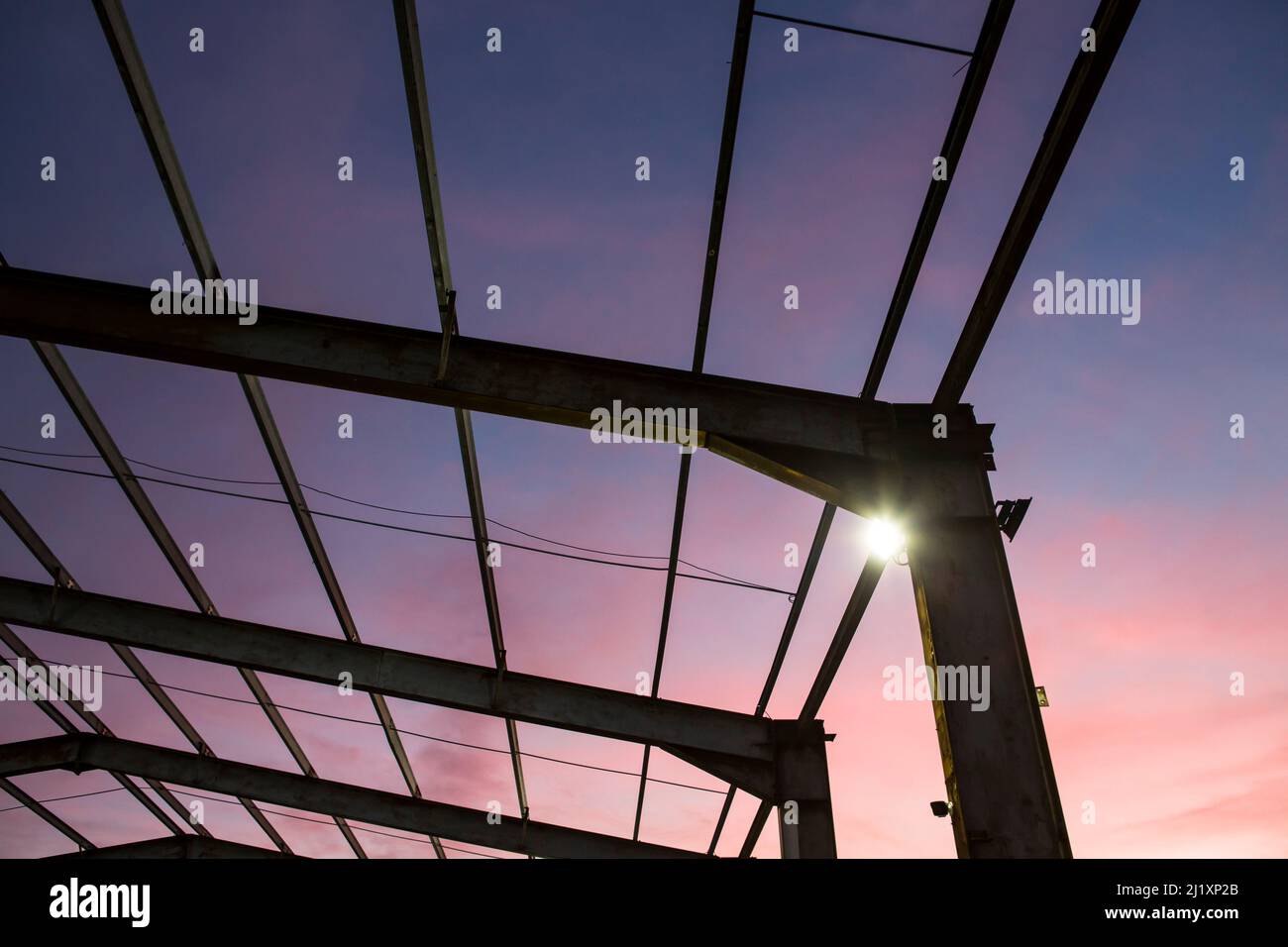 Blick nach oben durch ein Geflecht aus Stahlträgern. Das formte früher das Dach eines großen Gebäudes mit dem bunten Sonnenuntergang dahinter und kleinen hellen Flutlichtern an den aufrechten Pfosten. Stockfoto