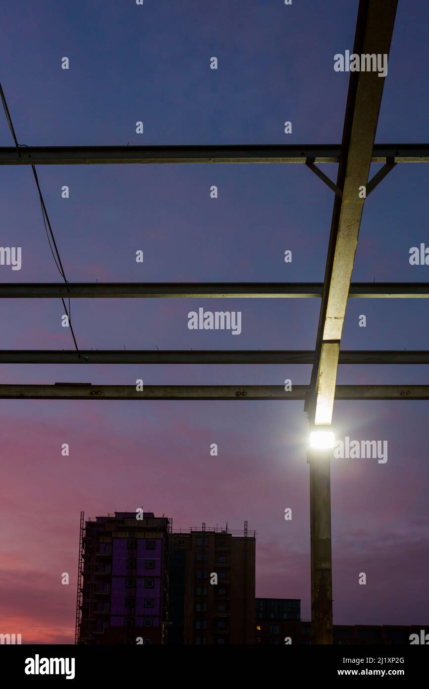 Blick nach oben durch ein Geflecht aus Stahlträgern. Das formte früher das Dach eines großen Gebäudes mit dem bunten Sonnenuntergang dahinter und kleinen hellen Flutlichtern an den aufrechten Pfosten. Stockfoto