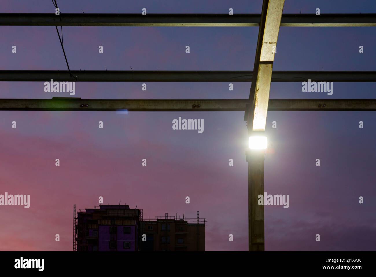 Blick nach oben durch ein Geflecht aus Stahlträgern. Das formte früher das Dach eines großen Gebäudes mit dem bunten Sonnenuntergang dahinter und kleinen hellen Flutlichtern an den aufrechten Pfosten. Stockfoto