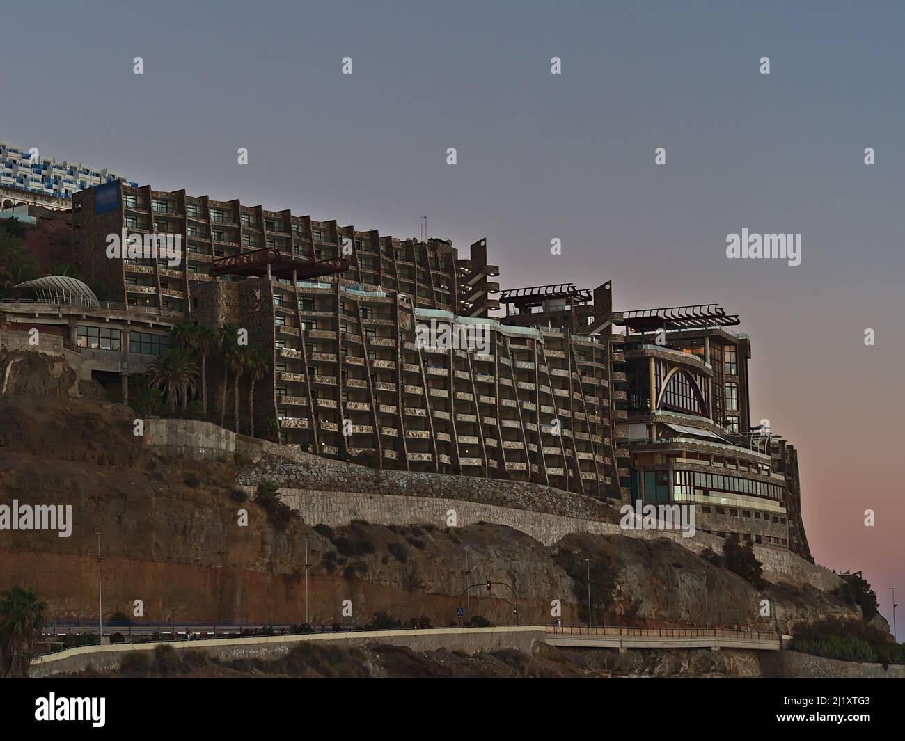 Blick auf großes hässliches Hotelgebäude an der Küste des Ferienortes Puerto Rico de Gran Canaria, Kanarische Inseln, Spanien am Abend mit Straße und Felsen. Stockfoto