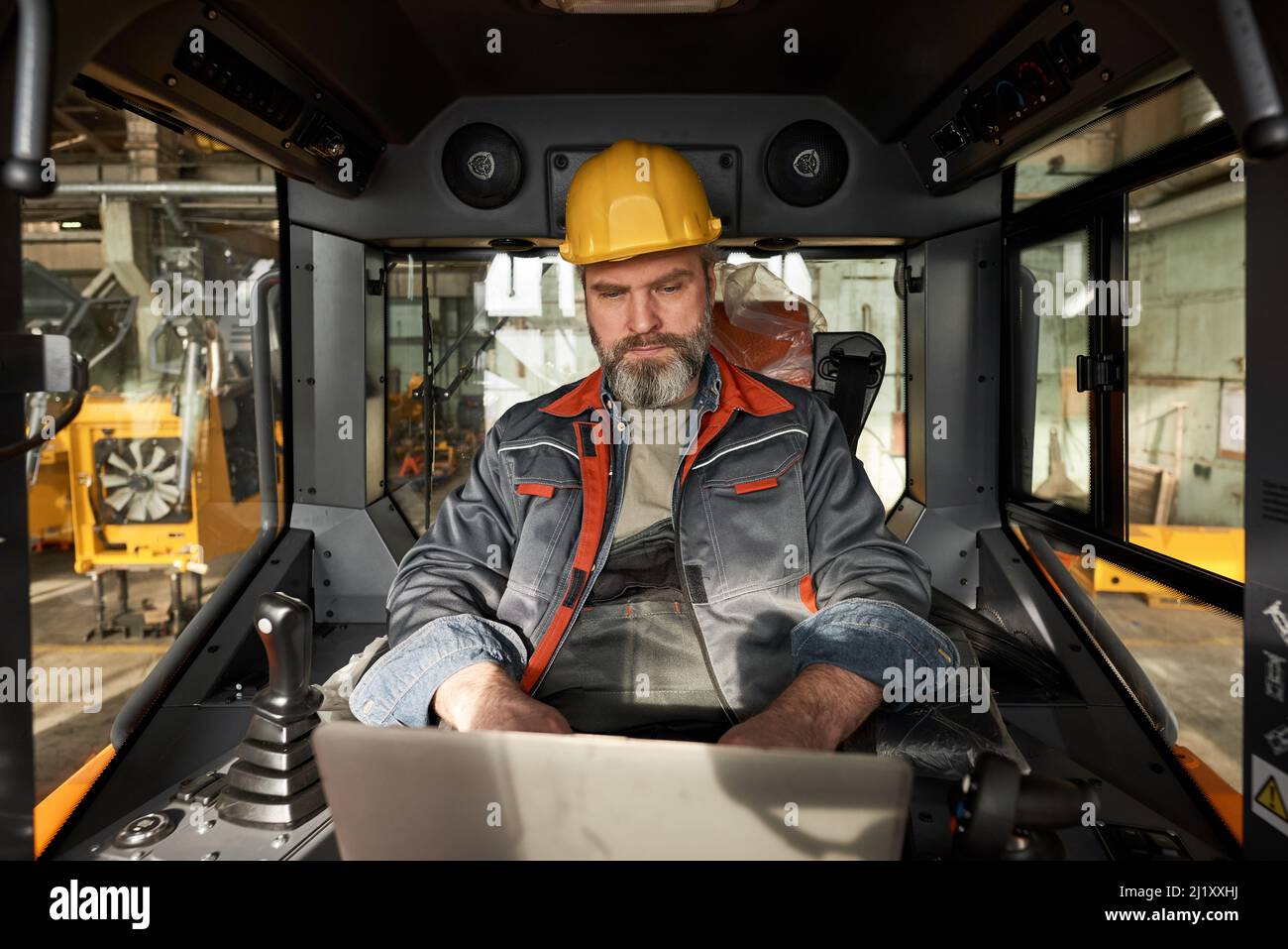 Reifer bärtiger Arbeiter in Uniform mit Laptop, während er während der Arbeiten auf der Baustelle in der Kabine der Maschine sitzt Stockfoto