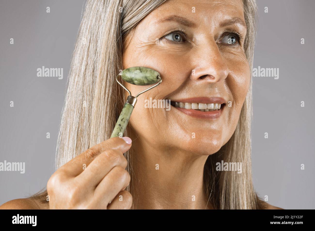 Porträt einer lächelnden reifen Frau mit natürlichen Jade Rolling Gerät für die Entgiftung der perfekten Haut über grauen Studio-Hintergrund. Schönheitstechnik der täglichen weiblichen Routine. Stockfoto