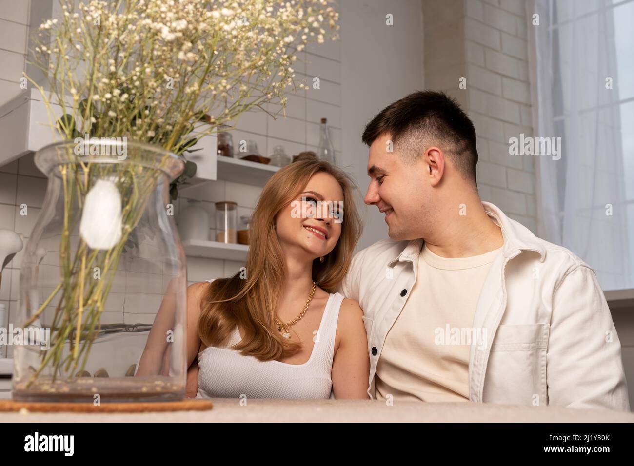 Glückliches junges Paar, das mit einer Vase mit getrockneten Blumen am Küchentisch sitzt, umarmt, einander anschaut und lächelt. Zartes, romantisches Beziehungskonzept. Hochwertige Fotos Stockfoto