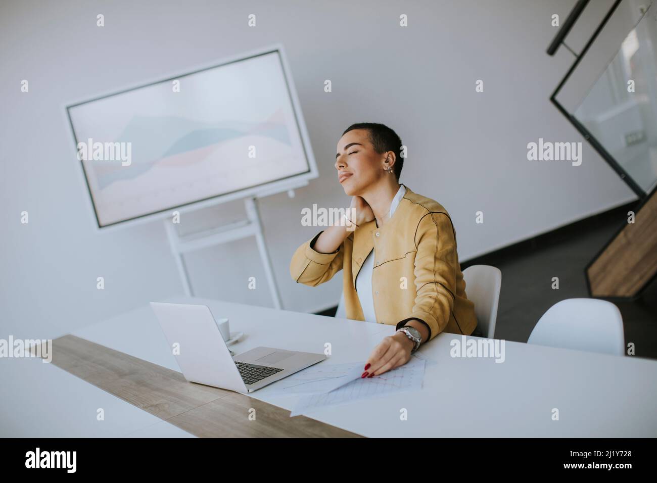 Müde junge Frau mit kurzen Haaren, die am Laptop arbeitet, während sie im Büro sitzt Stockfoto