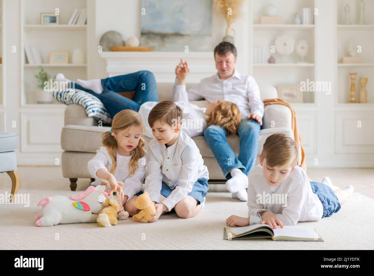 Gemütliche verträumte Eltern genießen Freizeit, Kinder sitzen auf dem Teppich und spielen Plüschtiere und lesen ein Buch, Mama Papa und drei kleine Teenager sind zu Hause. Familienbeziehungskonzept Stockfoto