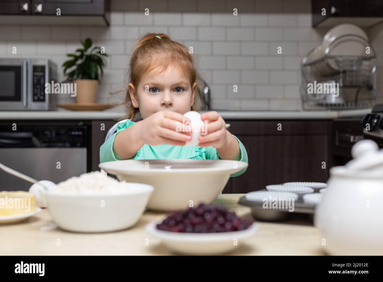 Kleines Mädchen, das in der Küche kocht. Kind sitzt am Tisch mit Essen. Kind zu Hause backen, Muffins kochen lernen, Eier in der Schüssel brechen Stockfoto