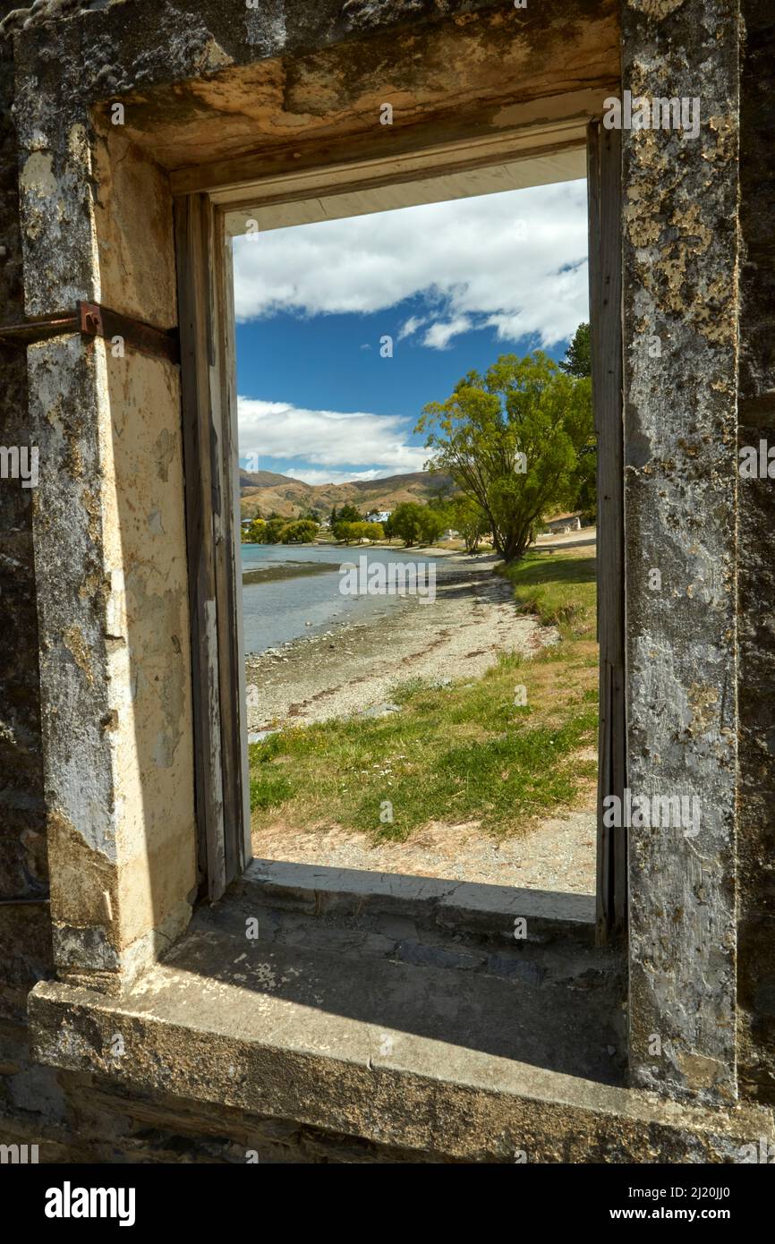 Blick durch das Fenster eines verroyten Gebäudes auf den Lake Dunstan und Old Cromwell Town, Cromwell, Central Otago, South Island, Neuseeland Stockfoto