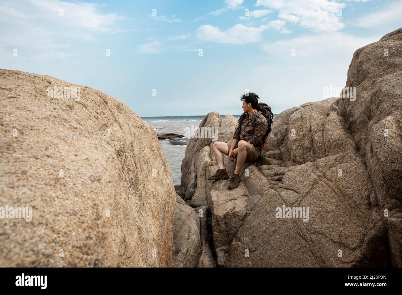 Junger Mann im Freiengehen, der auf einem Strandfelsen sitzt und die Landschaft betrachtet - Stock Photo Stockfoto