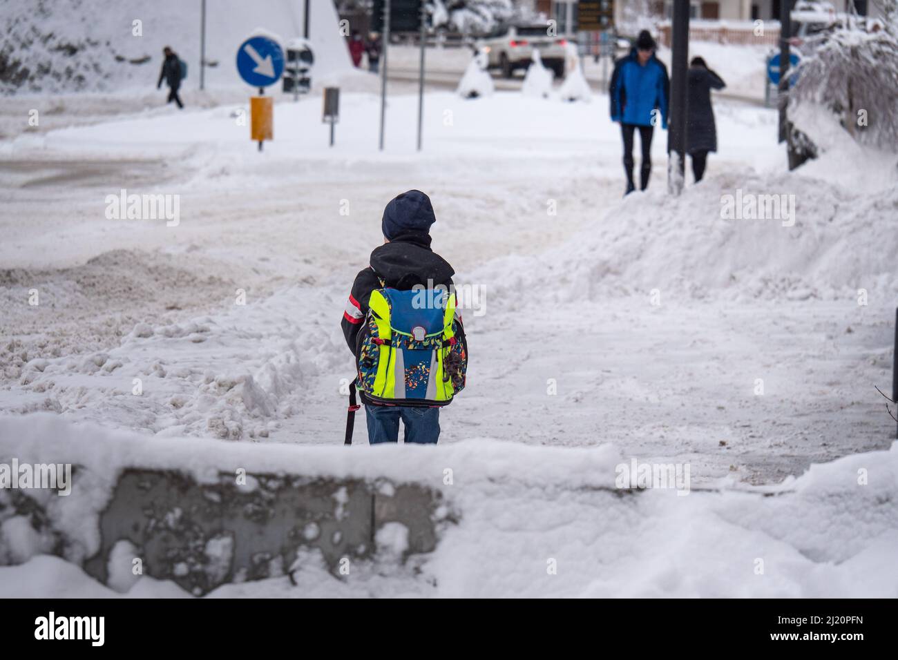 Kind, das etwa 7 Jahre alt ist und an einem kalten, verschneiten Wintertag eine verschneite Straße mit seiner Schultasche über der Schulter hinuntergeht. Stockfoto