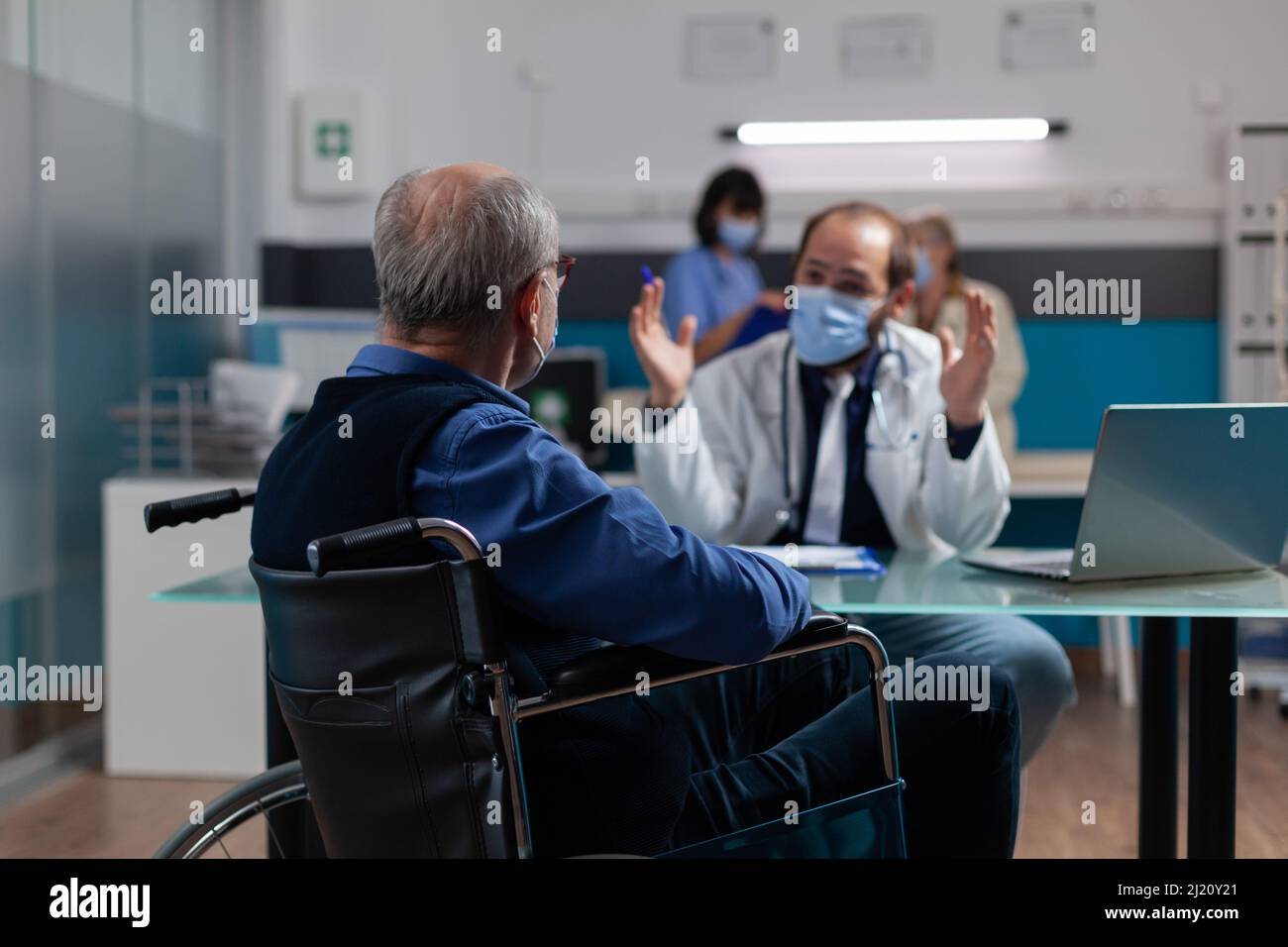 Fachgespräch mit Patient im Rollstuhl bei Termin zur Untersuchung während einer Pandemie. Ein Arzt und ein älterer Mann mit Gesichtsmaske machen eine Untersuchung und geben dem Patienten mit körperlicher Behinderung Ratschläge Stockfoto