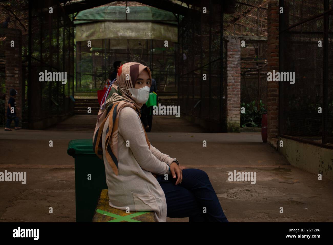 Schöne asiatische Mädchen, trägt eine Gesichtsmaske sitzen, in einem hellbraunen Kleid sitzen auf einem Stuhl im Gang Stockfoto