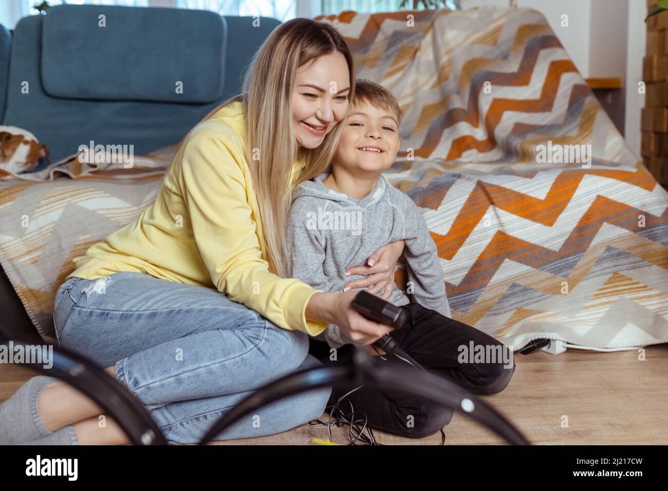 Mama und kleiner Sohn spielen zu Hause auf dem Teppich Rennen, haben Spaß und umarmen sich. Alleinerziehende Mutter zieht ihren Sohn mit Autos auf Stockfoto