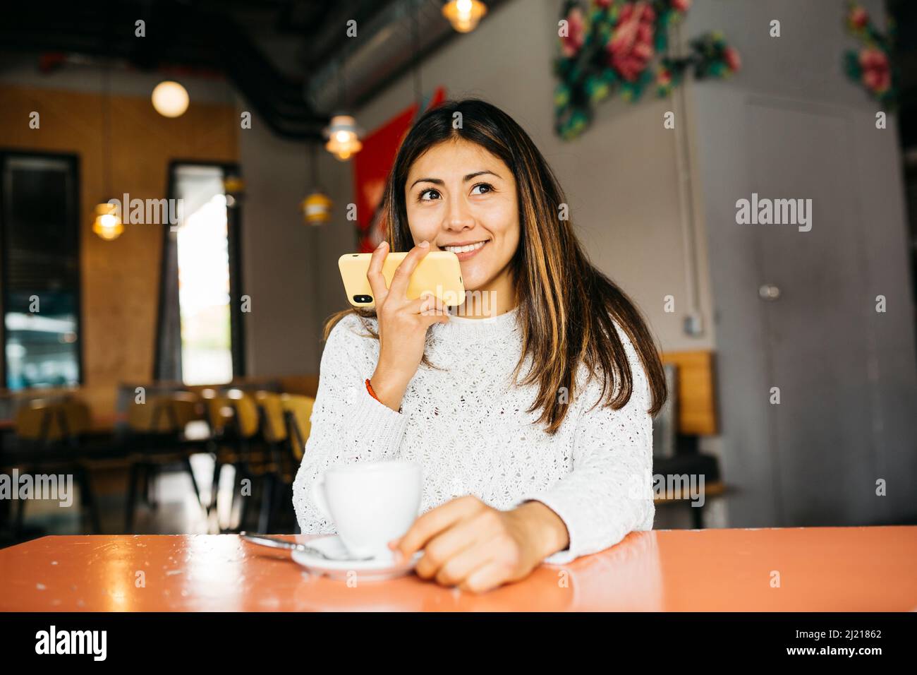 Porträt einer jungen lächelnden Frau, die in einem Café ein Telefon benutzt, während sie Kaffee trinkt Stockfoto